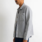 A man shown in profile wearing a light gray corduroy utility jacket over a dark striped shirt and dark blue jeans, standing against a plain white studio background.