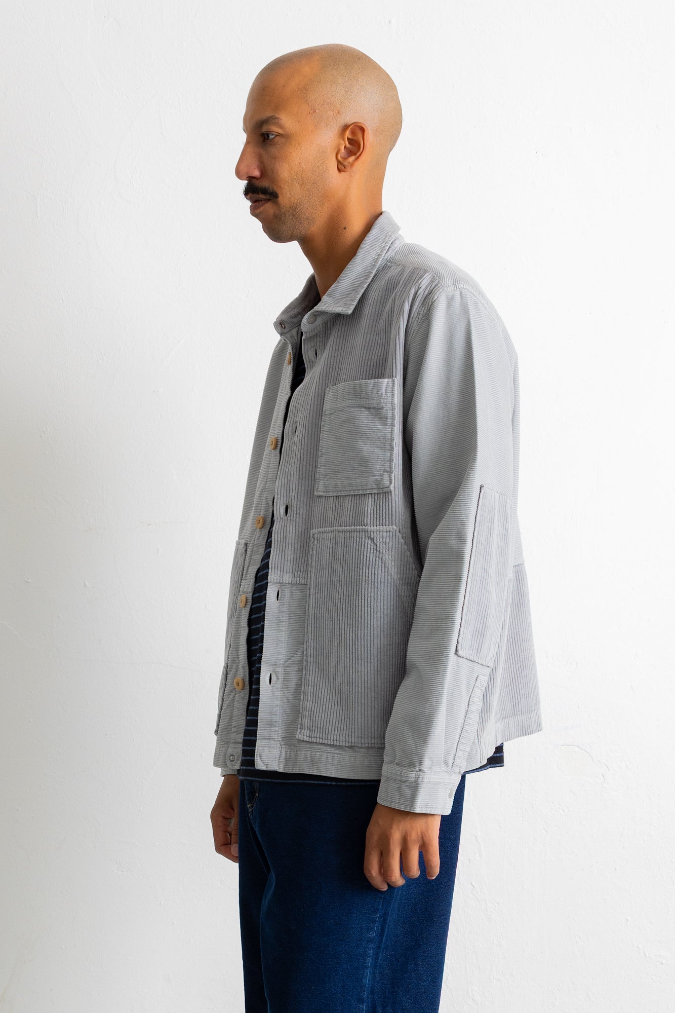 A man shown in profile wearing a light gray corduroy utility jacket over a dark striped shirt and dark blue jeans, standing against a plain white studio background.