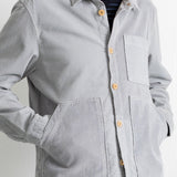 A man wearing a light gray corduroy button-up shirt jacket with a chest pocket and large front patch pockets, paired with a dark striped t-shirt and blue jeans.