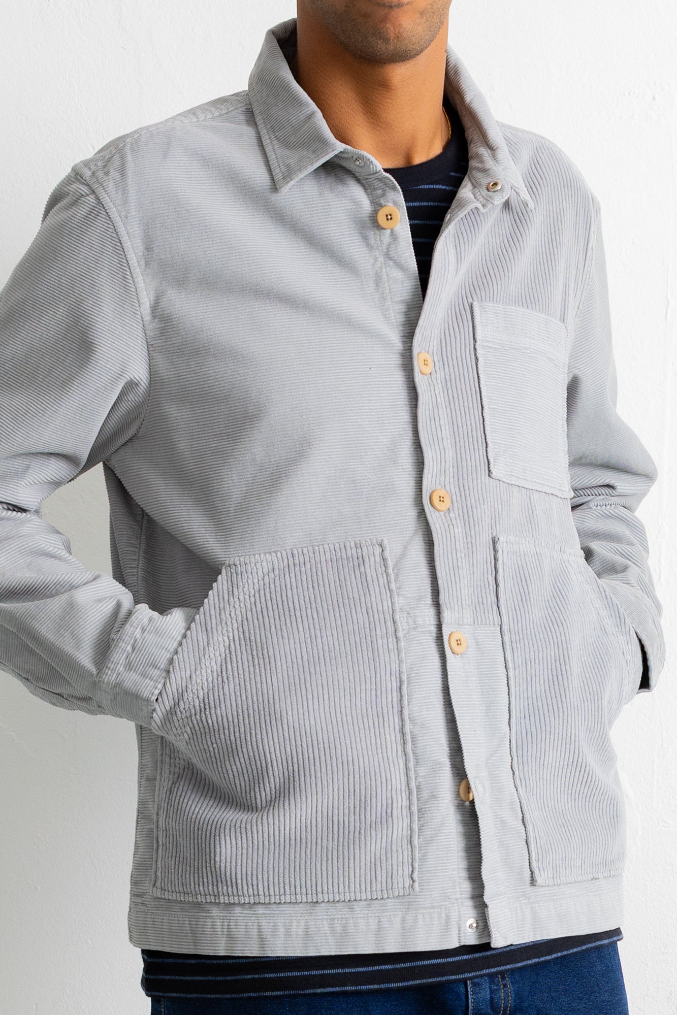 A man wearing a light gray corduroy button-up shirt jacket with a chest pocket and large front patch pockets, paired with a dark striped t-shirt and blue jeans.