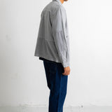 A man in profile wearing a light gray patchwork overshirt, dark blue jeans, and black canvas sneakers, standing against a white studio wall.