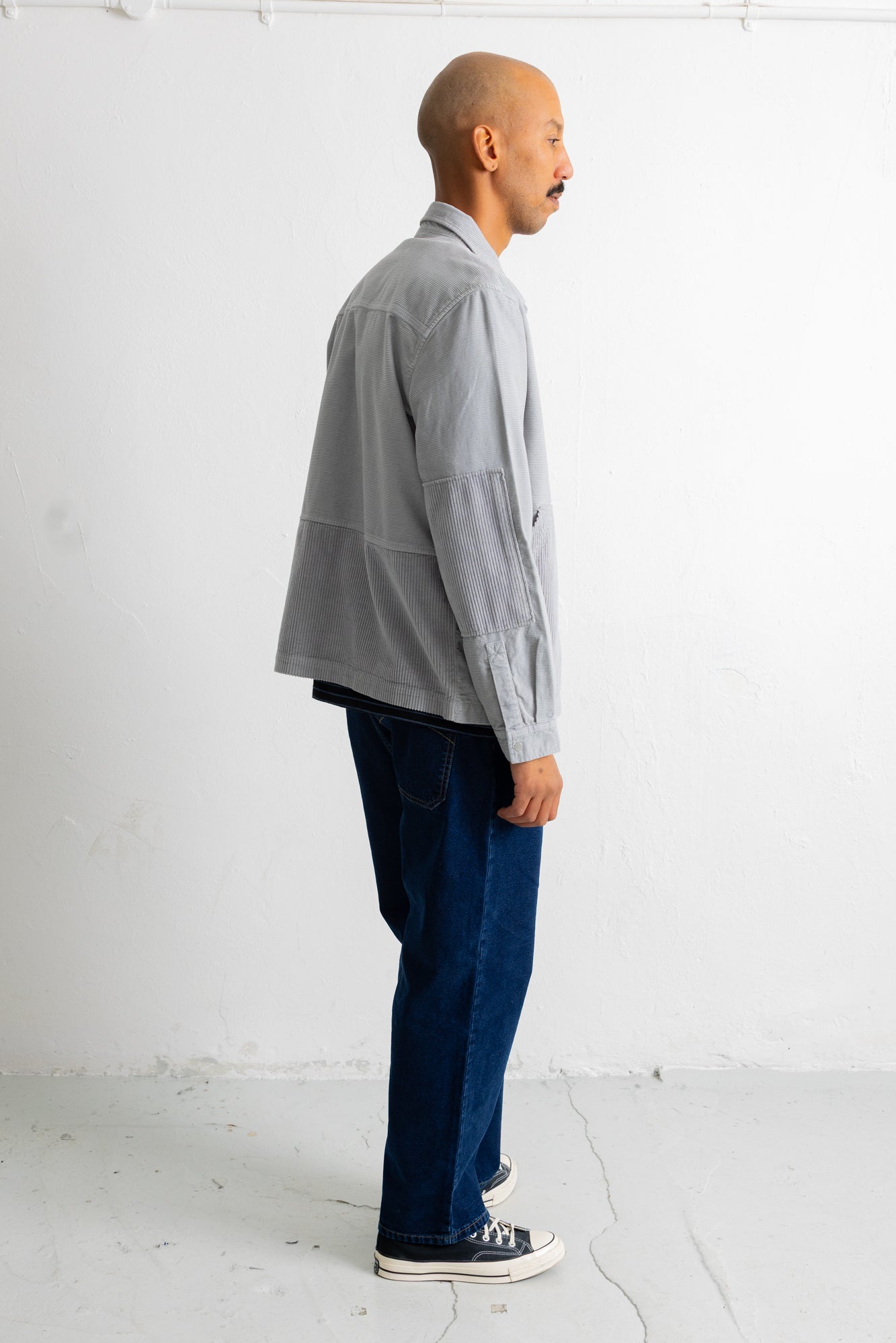 A man in profile wearing a light gray patchwork overshirt, dark blue jeans, and black canvas sneakers, standing against a white studio wall.