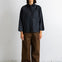 A model stands against a white wall wearing a dark navy textured long-sleeve button-up shirt and wide-leg brown trousers.