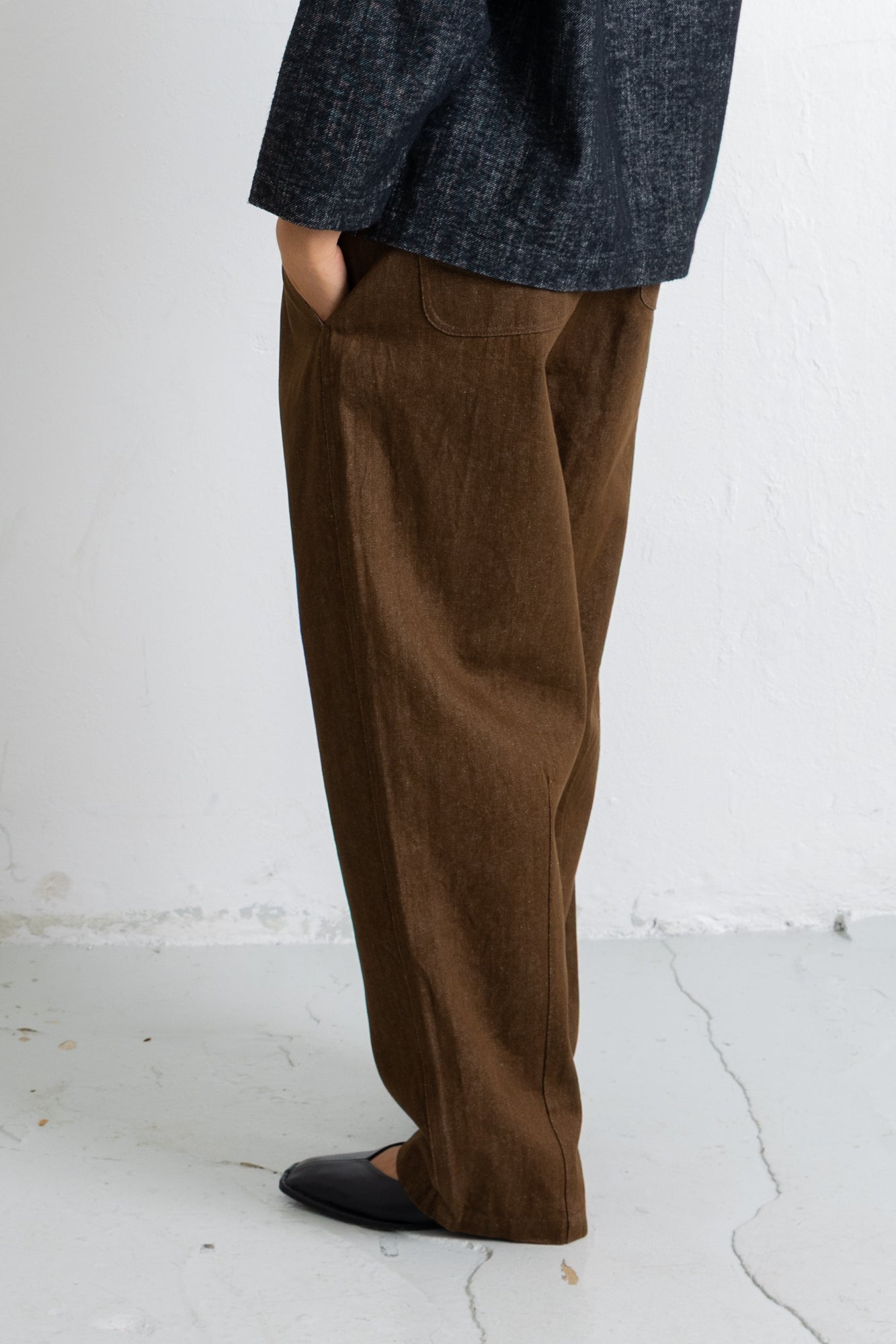 A person shown from the waist down wearing loose brown corduroy trousers, a dark textured blazer, and black shoes, standing against a white wall.