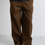 A person wearing loose brown wide-leg trousers, a dark charcoal textured jacket, and black pointed flats, standing against a light gray wall and concrete floor.
