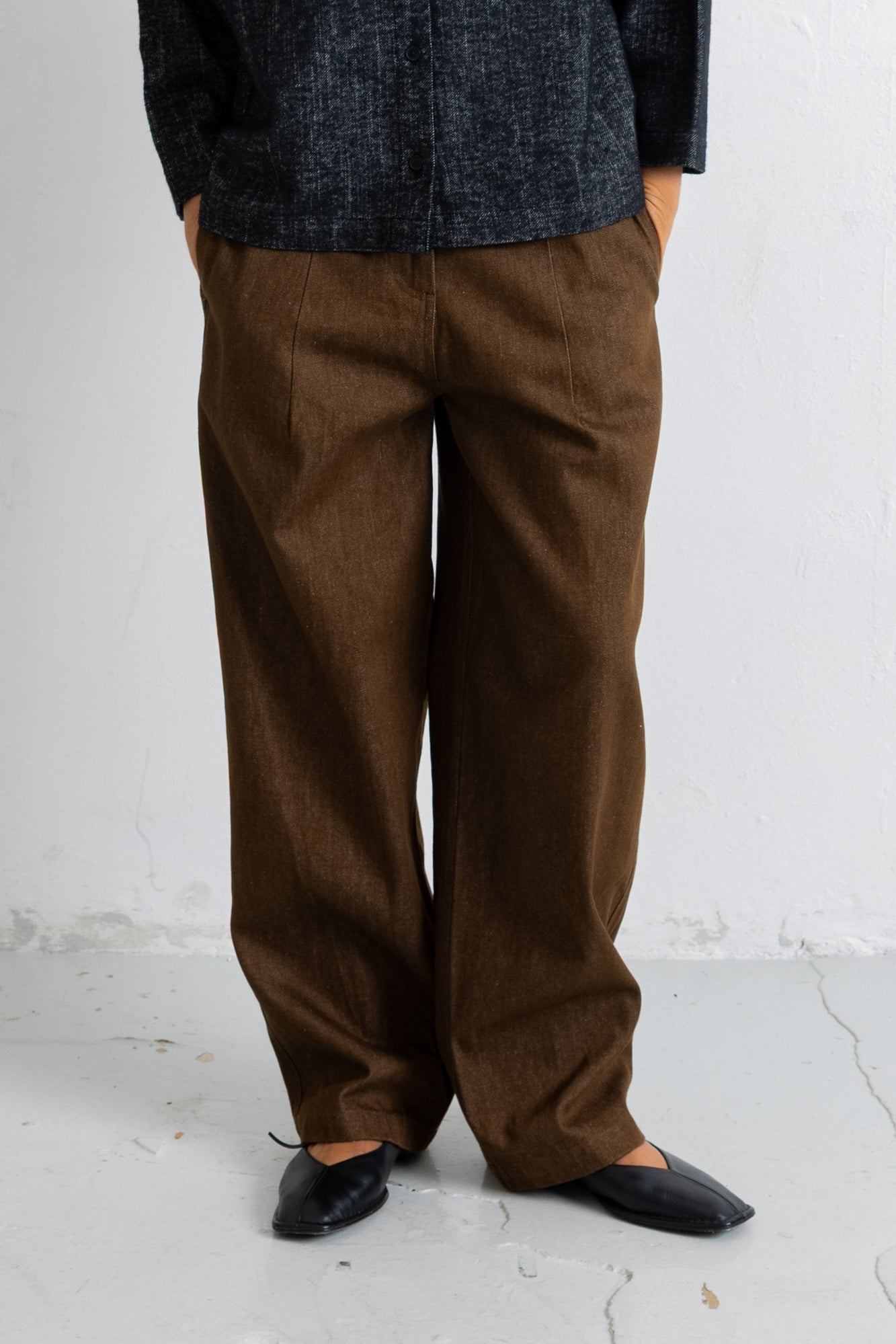A person wearing loose brown wide-leg trousers, a dark charcoal textured jacket, and black pointed flats, standing against a light gray wall and concrete floor.