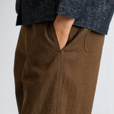 Close-up of a person wearing brown wide-leg trousers and a dark charcoal textured top, with one hand in the trouser pocket, against a plain light background.