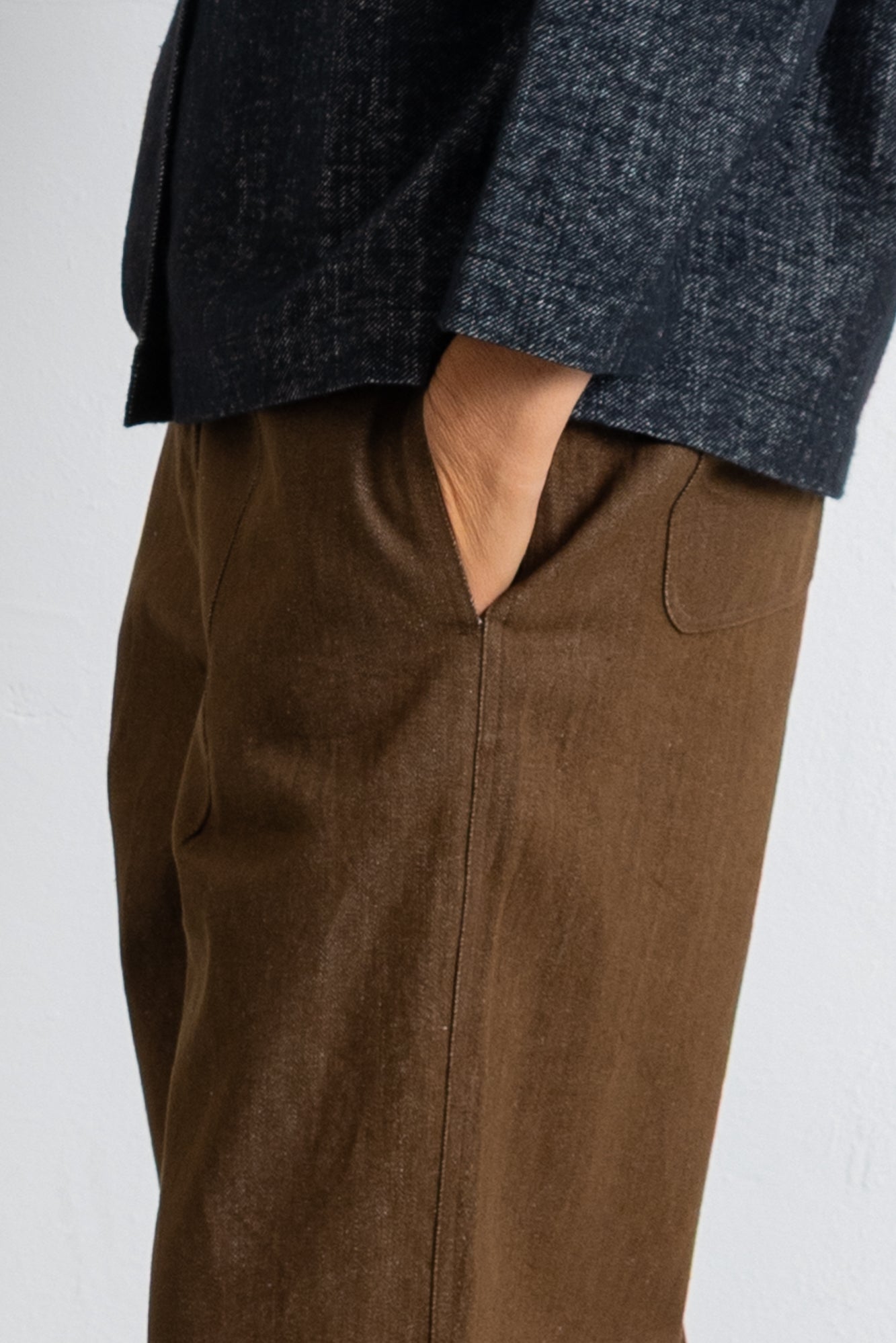 Close-up of a person wearing brown wide-leg trousers and a dark charcoal textured top, with one hand in the trouser pocket, against a plain light background.