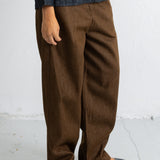 A person wearing loose brown wide-leg trousers with a dark charcoal knit top and black pointed flats, standing against a white wall.