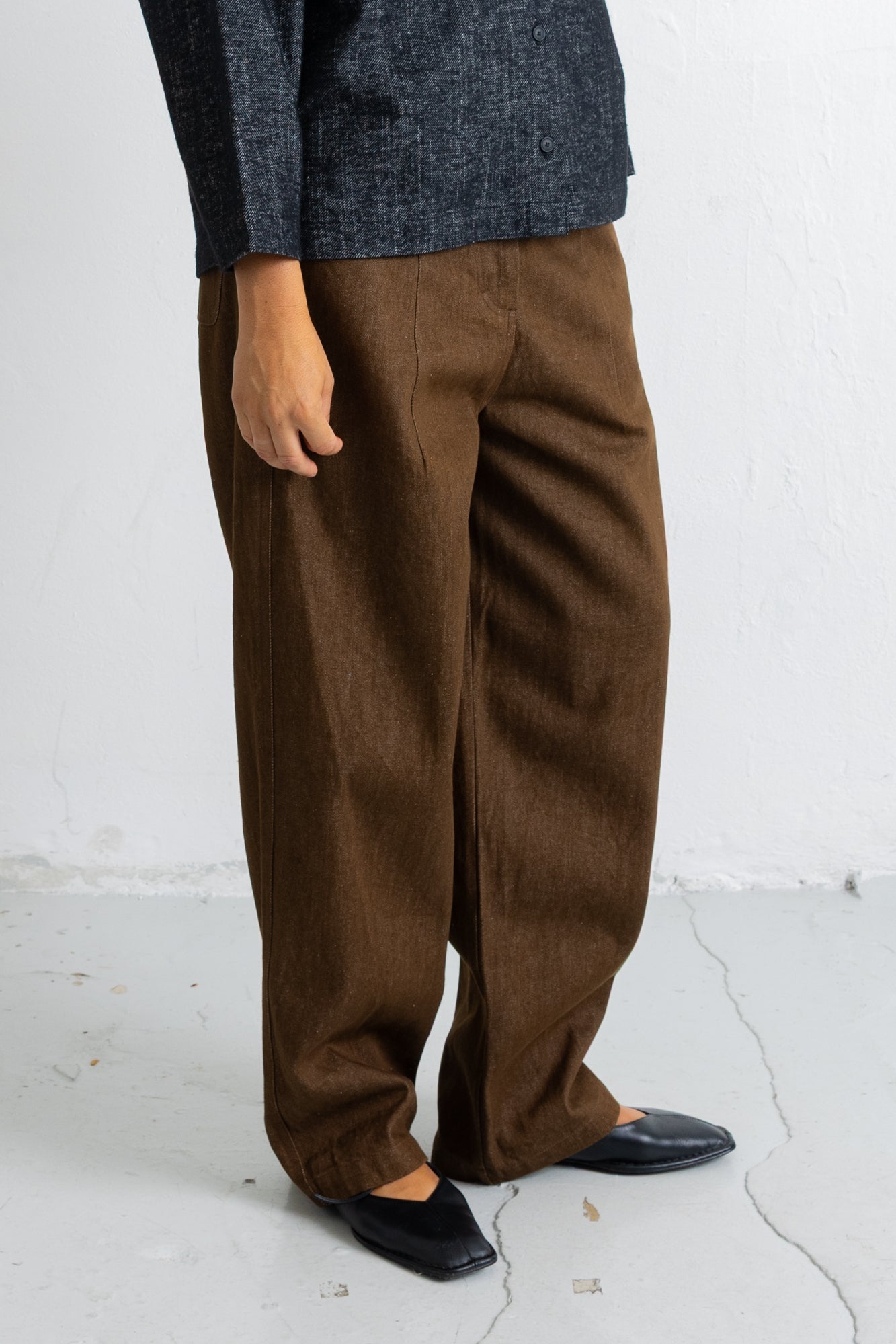 A person wearing loose brown wide-leg trousers with a dark charcoal knit top and black pointed flats, standing against a white wall.