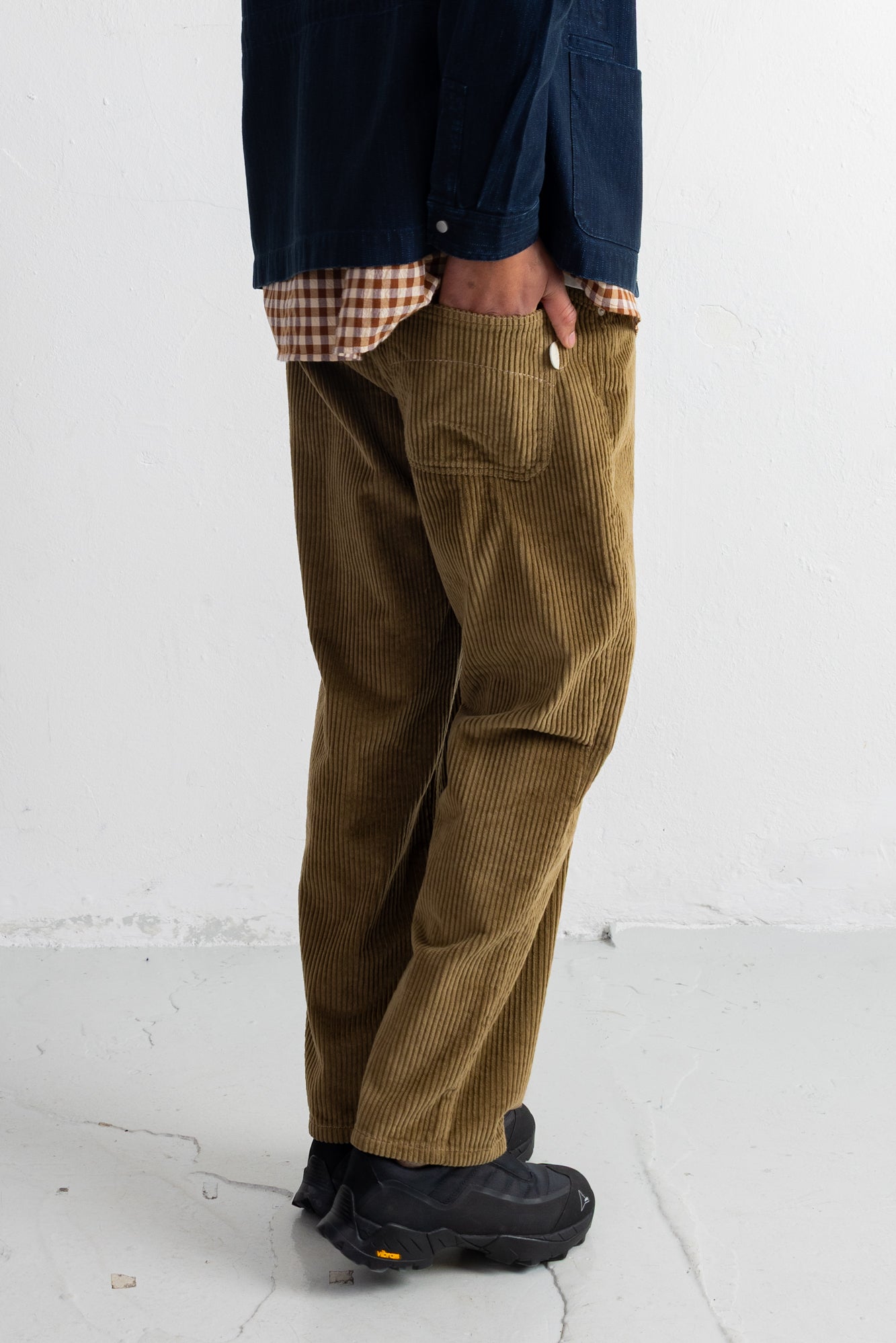A person wearing brown corduroy pants with a navy shirt and black shoes, standing against a white wall.