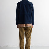 A man viewed from behind wearing a dark navy blue jacket, brown corduroy trousers, and black shoes, standing against a plain white studio wall.