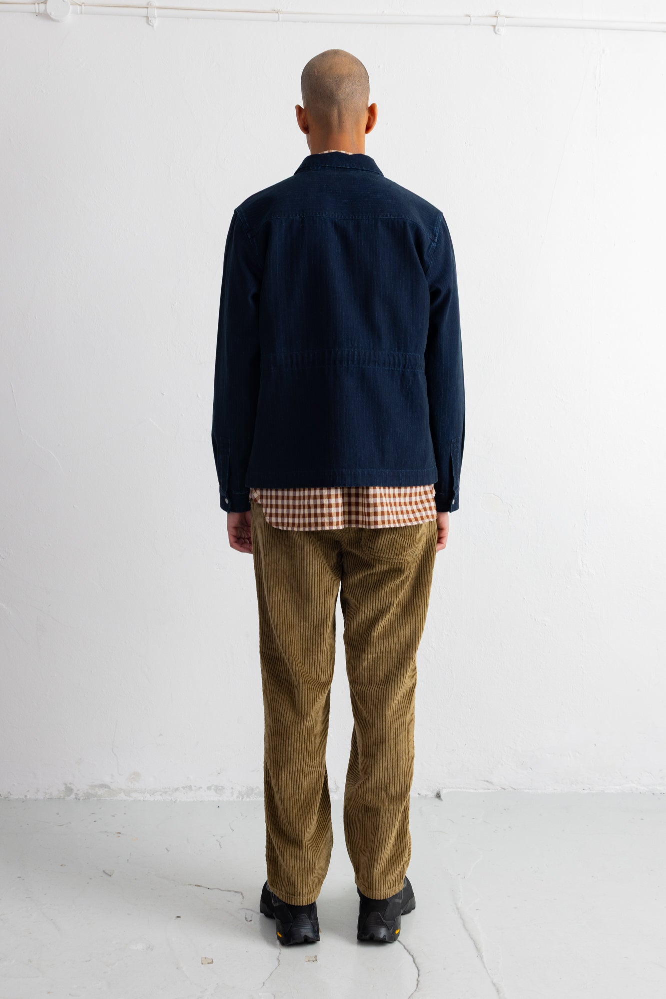 A man viewed from behind wearing a dark navy blue jacket, brown corduroy trousers, and black shoes, standing against a plain white studio wall.