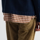 Close-up of a person wearing a navy overshirt over a brown and beige gingham shirt, paired with olive corduroy trousers, against a plain white wall.