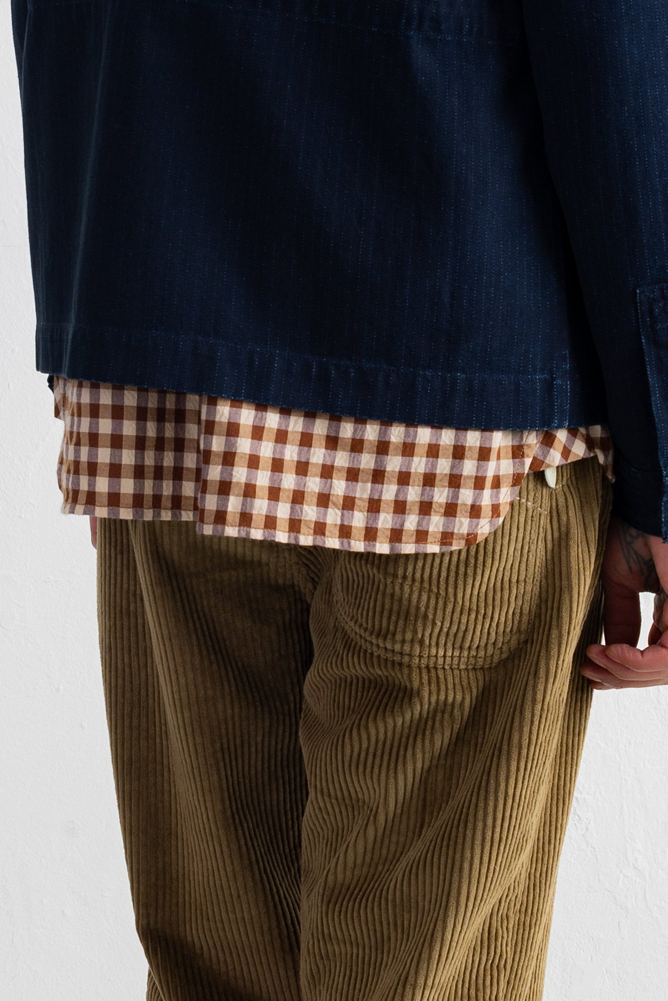 Close-up of a person wearing a navy overshirt over a brown and beige gingham shirt, paired with olive corduroy trousers, against a plain white wall.
