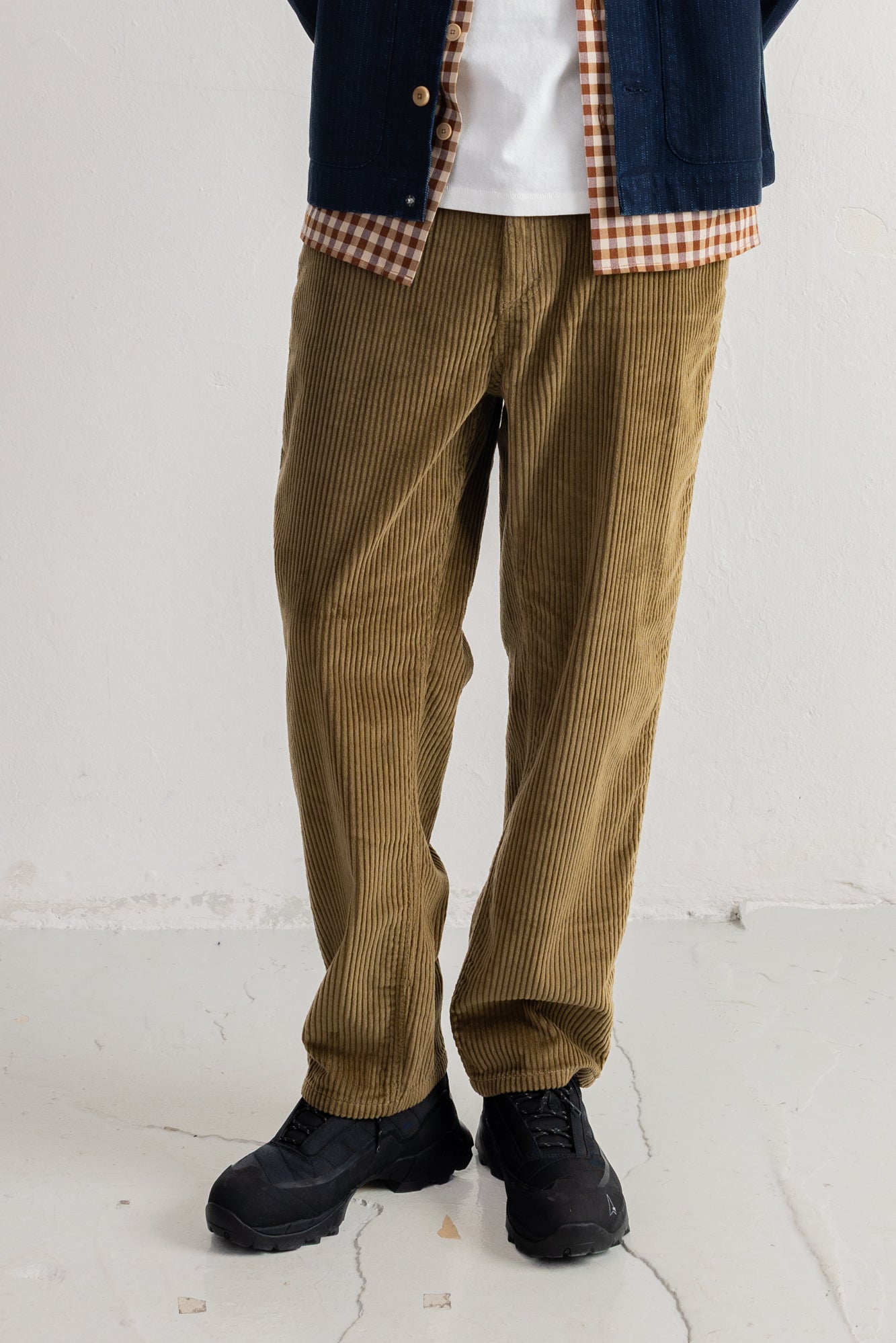 A person wearing loose brown corduroy pants, a navy cardigan over a white shirt, and black shoes, standing against a plain light wall.