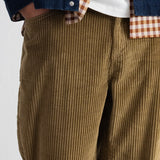 Close-up of a person wearing olive-brown corduroy pants, layered with a white t-shirt, a brown-and-white gingham shirt, and a dark blue denim jacket.