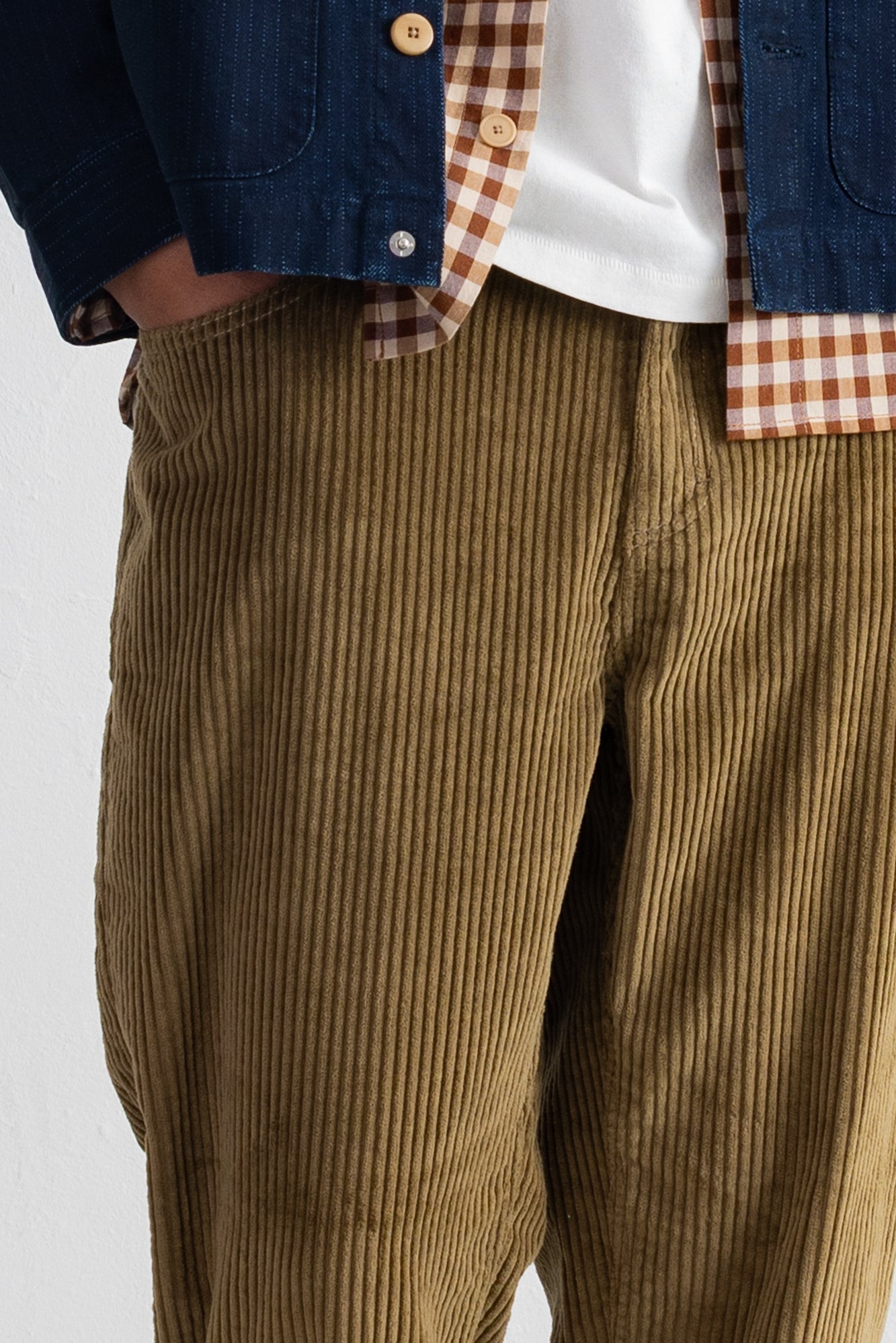 Close-up of a person wearing olive-brown corduroy pants, layered with a white t-shirt, a brown-and-white gingham shirt, and a dark blue denim jacket.