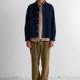 A man stands against a white studio wall wearing a dark navy overshirt over a brown and beige gingham shirt, olive-brown corduroy pants, and black shoes.