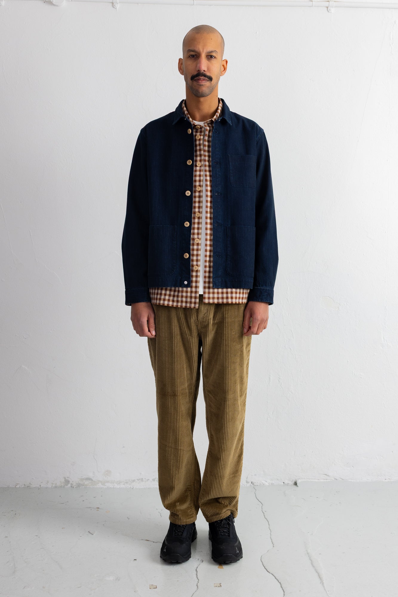 A man stands against a white studio wall wearing a dark navy overshirt over a brown and beige gingham shirt, olive-brown corduroy pants, and black shoes.
