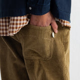 Close-up of a person wearing olive-brown corduroy pants with a hand in the pocket, layered over a brown-and-white gingham shirt and a dark blue jacket.