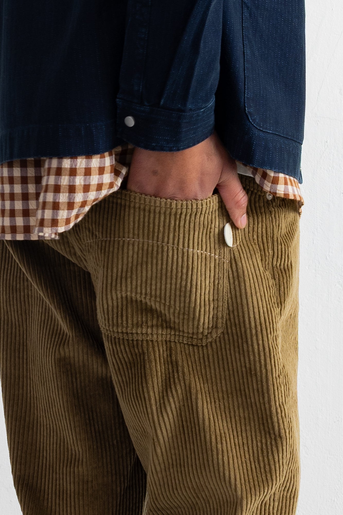 Close-up of a person wearing olive-brown corduroy pants with a hand in the pocket, layered over a brown-and-white gingham shirt and a dark blue jacket.