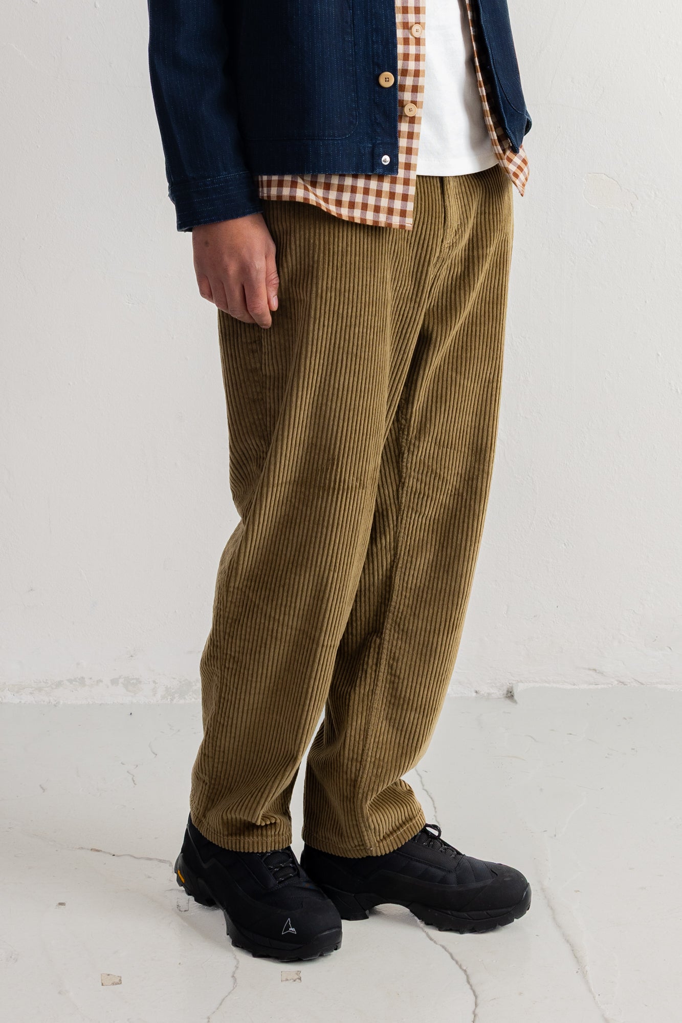 A person wearing loose olive-brown corduroy pants, a navy jacket over a white and tan check shirt, and black shoes, standing against a plain off-white wall.