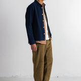 A man stands in profile wearing a dark navy overshirt over a white and brown gingham shirt, paired with olive brown corduroy trousers and black shoes, against a plain white studio wall.