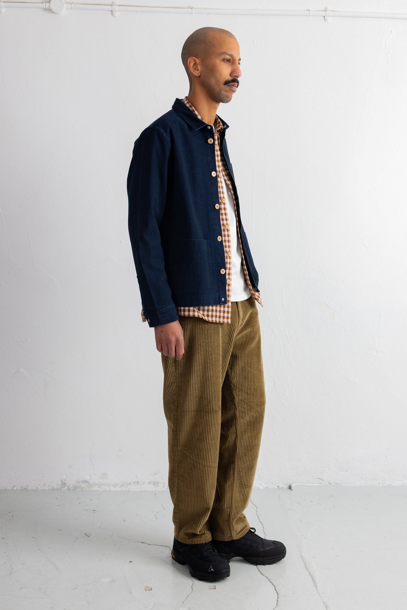 A man stands in profile wearing a dark navy overshirt over a white and brown gingham shirt, paired with olive brown corduroy trousers and black shoes, against a plain white studio wall.