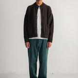 A man stands against a plain white studio wall wearing a dark brown button-front cardigan over a black-and-white striped shirt, with teal-green pants and black shoes.