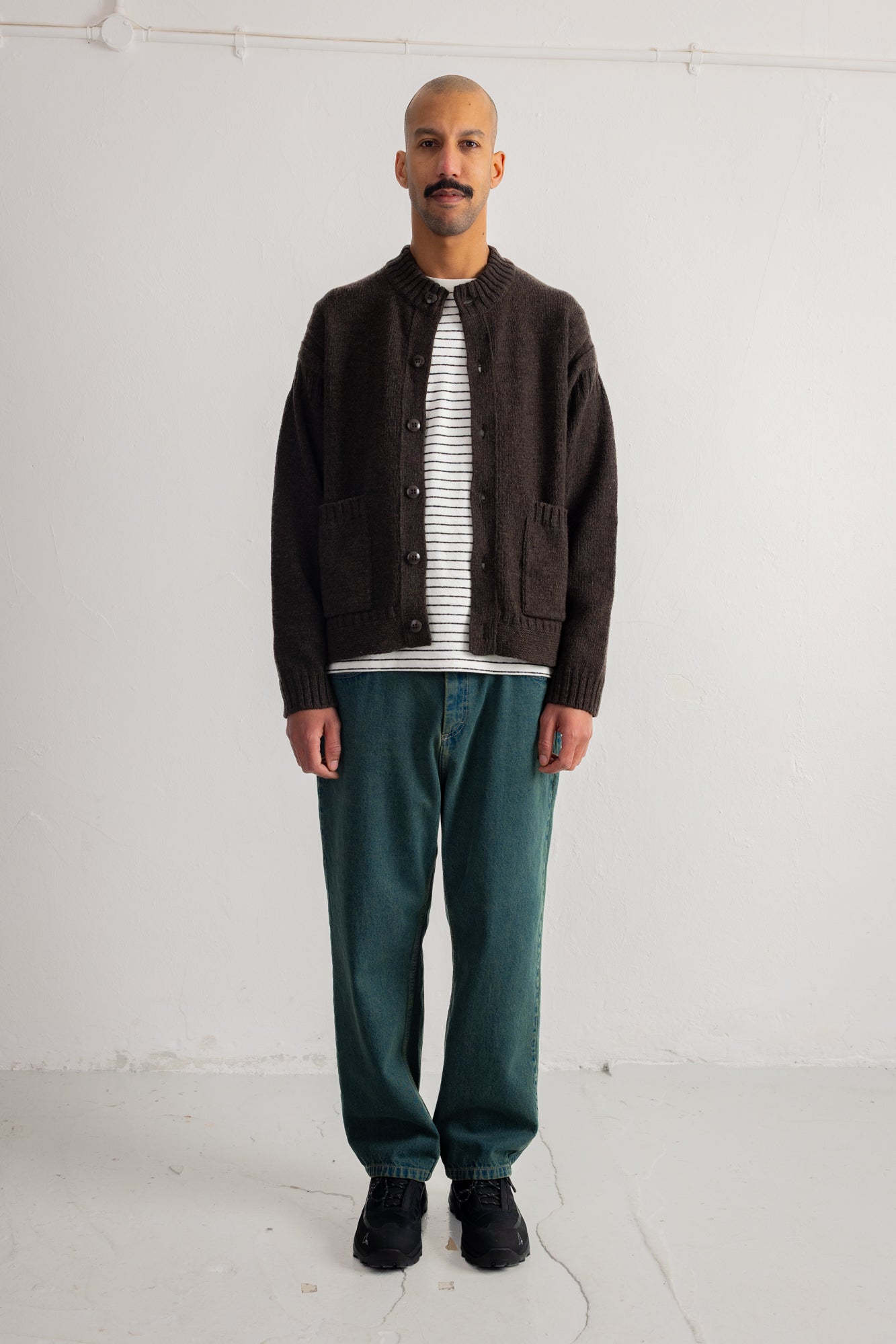 A man stands against a plain white studio wall wearing a dark brown button-front cardigan over a black-and-white striped shirt, with teal-green pants and black shoes.
