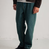 A person wearing dark teal-green straight-leg jeans, a dark brown cardigan over a white striped shirt, and black sneakers, standing against a white wall.