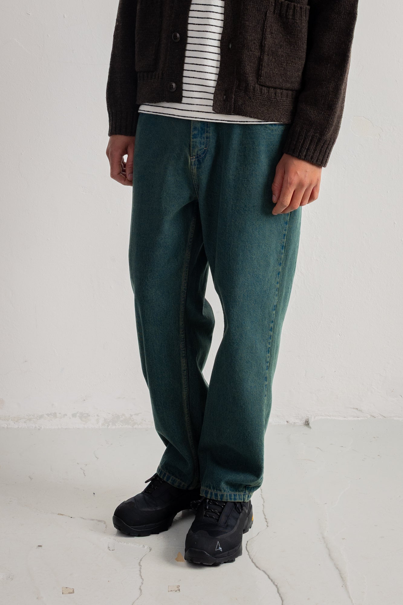 A person wearing dark teal-green straight-leg jeans, a dark brown cardigan over a white striped shirt, and black sneakers, standing against a white wall.