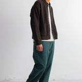 A man wearing a dark brown knit cardigan over a black and white striped shirt, paired with teal green trousers and black shoes, standing against a plain white studio background.
