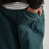 Close-up of a person wearing dark teal blue denim jeans with a hand in the back pocket, styled with a striped shirt and a dark brown knit sweater.