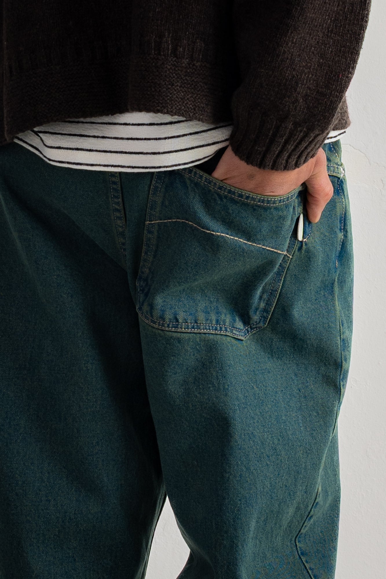 Close-up of a person wearing dark teal blue denim jeans with a hand in the back pocket, styled with a striped shirt and a dark brown knit sweater.