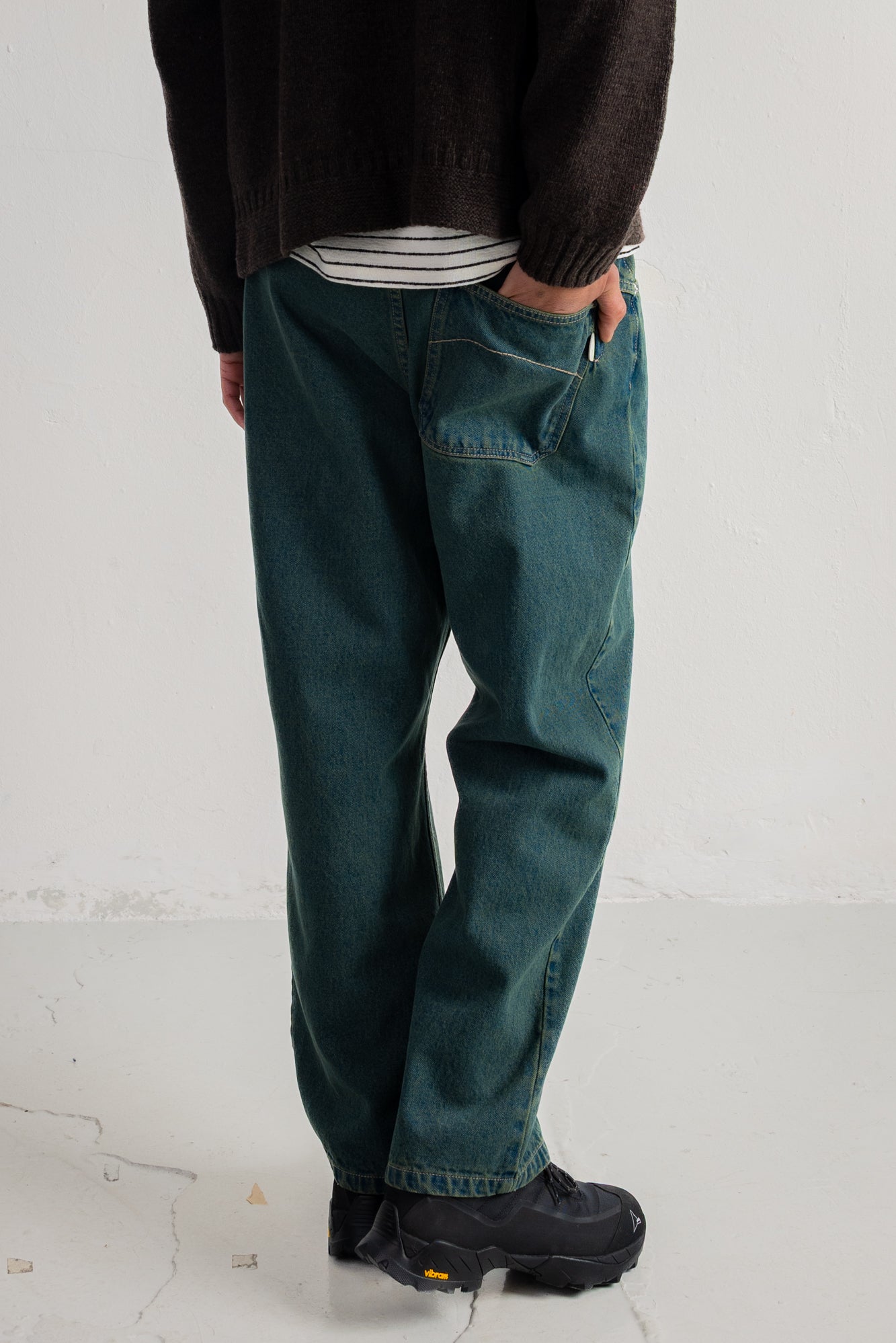 A person shown from the waist down wearing dark teal blue denim jeans, a black knit sweater, a striped shirt, and black shoes, standing against a white wall.