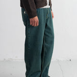 A person wearing loose teal-green corduroy pants, a dark brown cardigan over a white striped shirt, and black shoes, standing against a white wall.