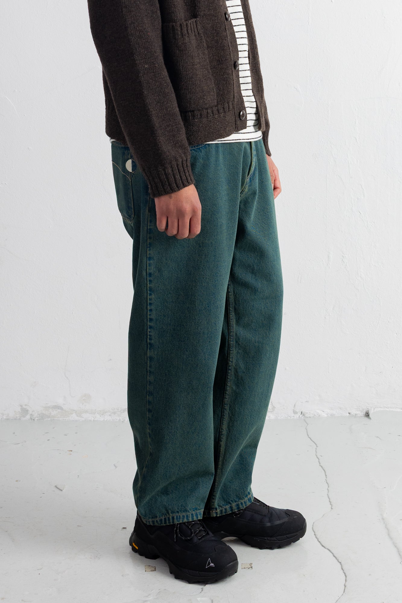A person wearing loose teal-green corduroy pants, a dark brown cardigan over a white striped shirt, and black shoes, standing against a white wall.
