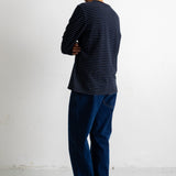 A man standing with his back to the camera, wearing a navy blue long-sleeve shirt with thin white horizontal stripes, dark blue jeans, and black canvas sneakers with white soles, set against a plain white wall.