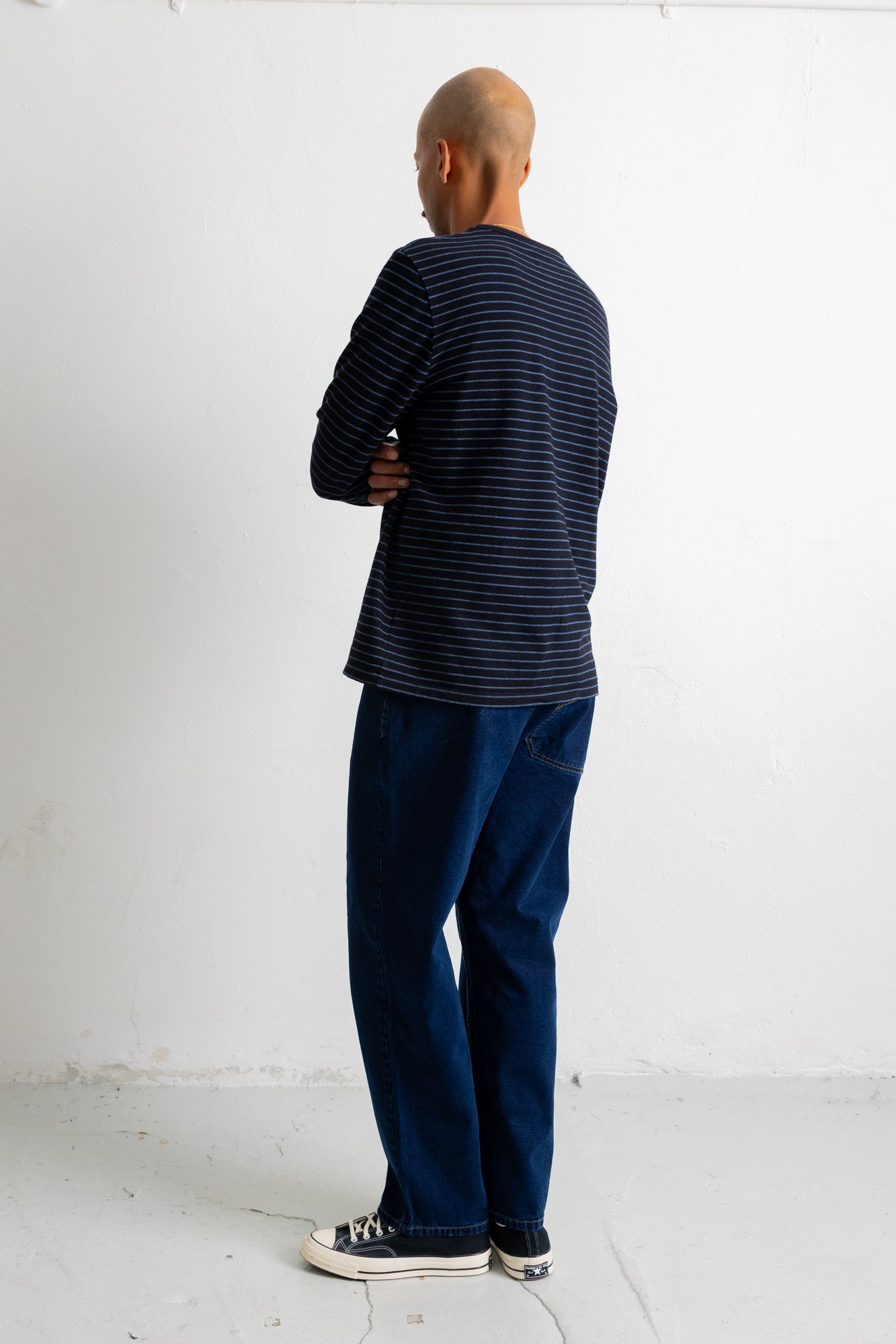 A man standing with his back to the camera, wearing a navy blue long-sleeve shirt with thin white horizontal stripes, dark blue jeans, and black canvas sneakers with white soles, set against a plain white wall.