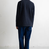 A man viewed from behind wearing a navy blue long-sleeve shirt with thin horizontal dark stripes, dark blue jeans, and black canvas sneakers, standing against a plain white wall.