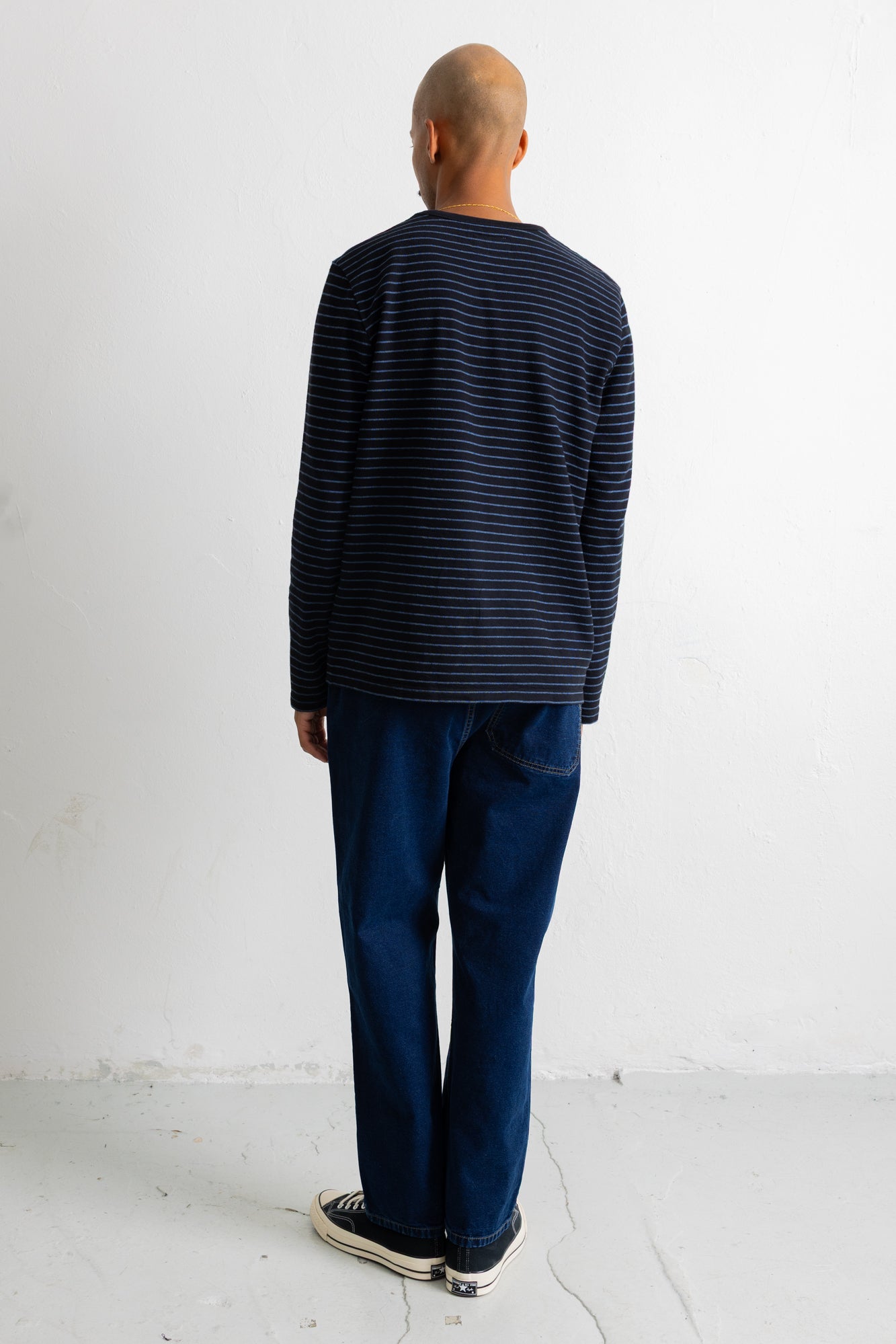 A man viewed from behind wearing a navy blue long-sleeve shirt with thin horizontal dark stripes, dark blue jeans, and black canvas sneakers, standing against a plain white wall.