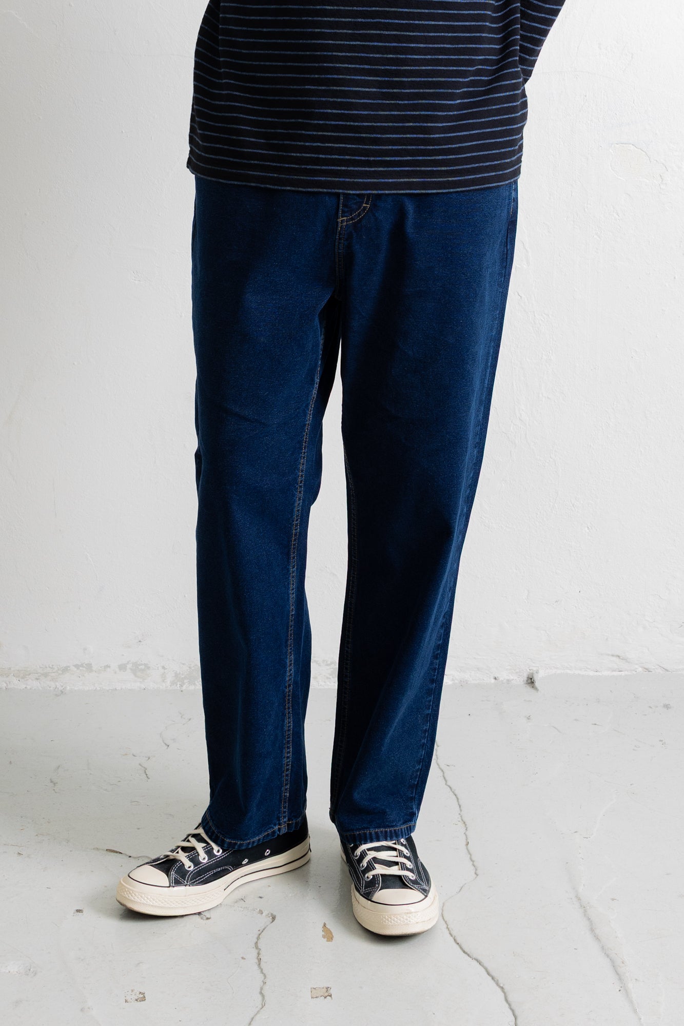 A person wearing dark blue straight-leg jeans, a navy and white striped t-shirt, and navy canvas sneakers with white soles, standing against a white wall.