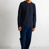 A man wearing a navy blue long-sleeve shirt with thin horizontal dark stripes, dark blue jeans, and black-and-white sneakers, standing against a plain white wall.
