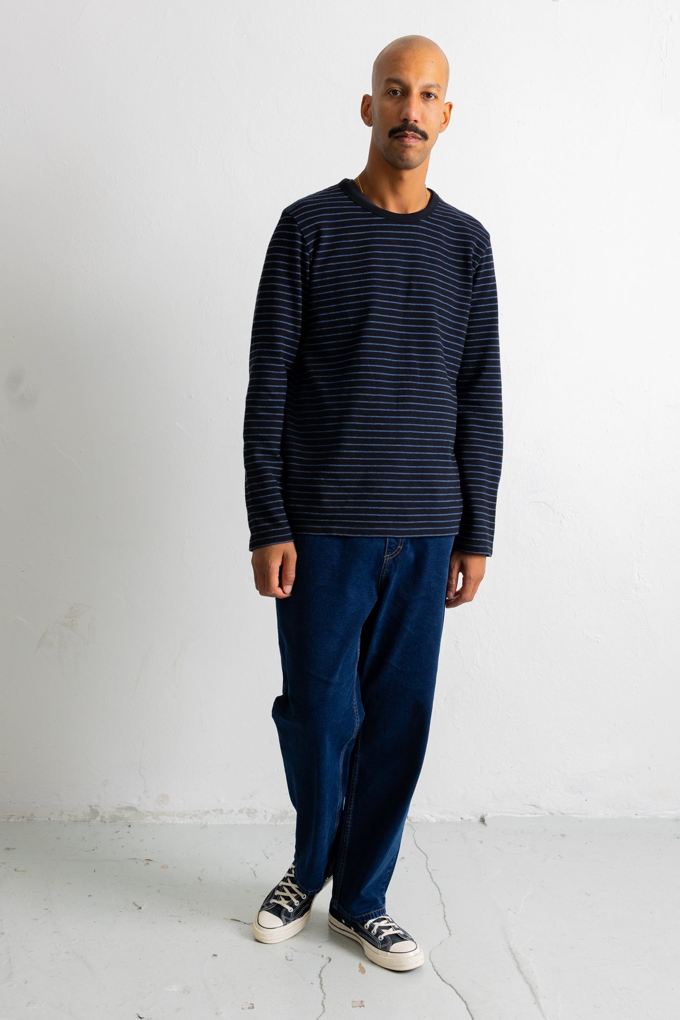 A man wearing a navy blue long-sleeve shirt with thin horizontal dark stripes, dark blue jeans, and black-and-white sneakers, standing against a plain white wall.