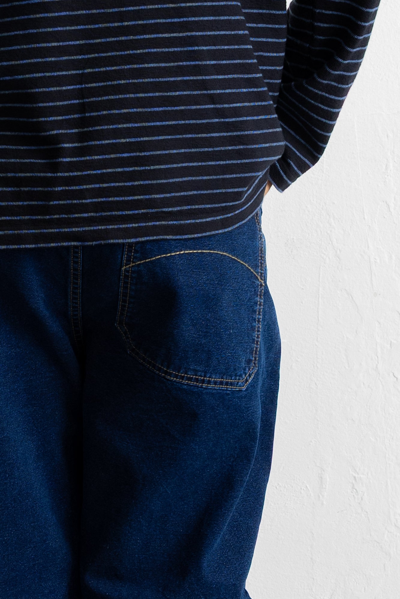 Close-up of a person wearing dark blue jeans with a back pocket and a navy striped top against a white wall.