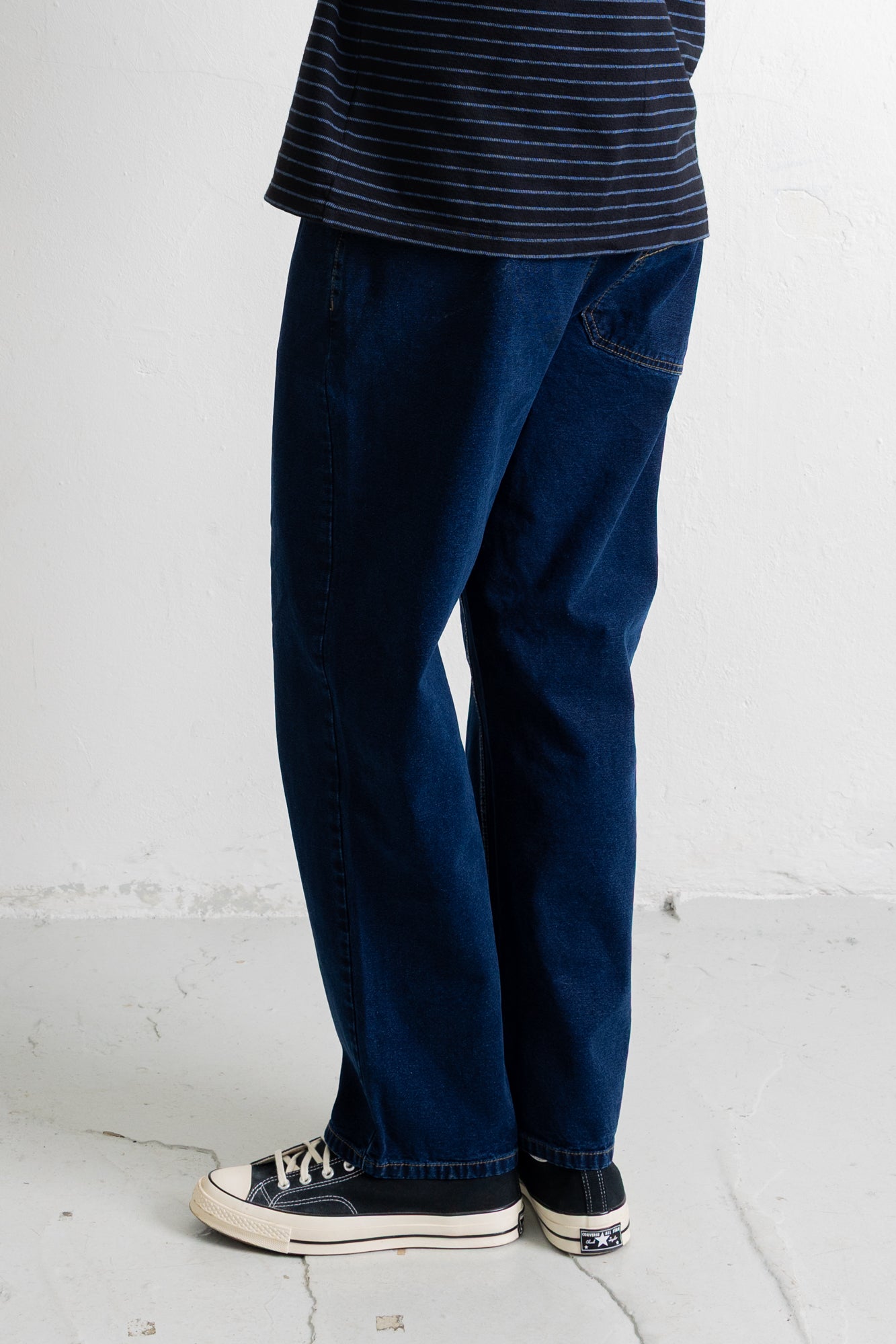 A person shown from the waist down wearing dark blue straight-leg jeans, a navy and white striped t-shirt, and black canvas sneakers with white soles, standing against a white wall.