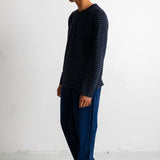 A man wearing a navy blue long-sleeve shirt with thin dark horizontal stripes, dark blue jeans, and white sneakers, standing in profile against a white studio wall.