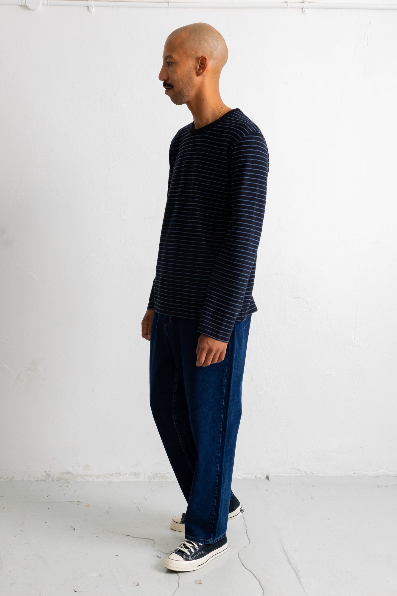 A man wearing a navy blue long-sleeve shirt with thin dark horizontal stripes, dark blue jeans, and white sneakers, standing in profile against a white studio wall.